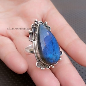 May include: A silver ring featuring a large, teardrop-shaped blue gemstone. The gemstone has a shimmering, iridescent quality, with flashes of blue and purple. The ring has an ornate, decorative setting with the text "SilverArtDesign925".