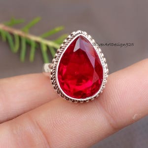 May include: A silver ring featuring a large, faceted, teardrop-shaped, bright red gemstone. The gemstone is set in a silver bezel with a twisted rope design around the edge. The text "SilverArtDesign925" is visible.
