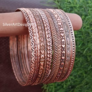 May include: A set of 12 copper bangles with various embossed patterns. The bangles are stacked on top of each other and are all different sizes.