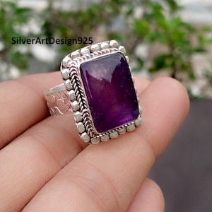 Può includere: Un anello d'argento con una grande pietra di ametista rettangolare viola scuro. L'anello ha un bordo decorativo di piccole perle d'argento e un design argentato intrecciato attorno alla pietra. La fascia ha un motivo strutturato. Il testo "SilverArtDesign925" è visibile.