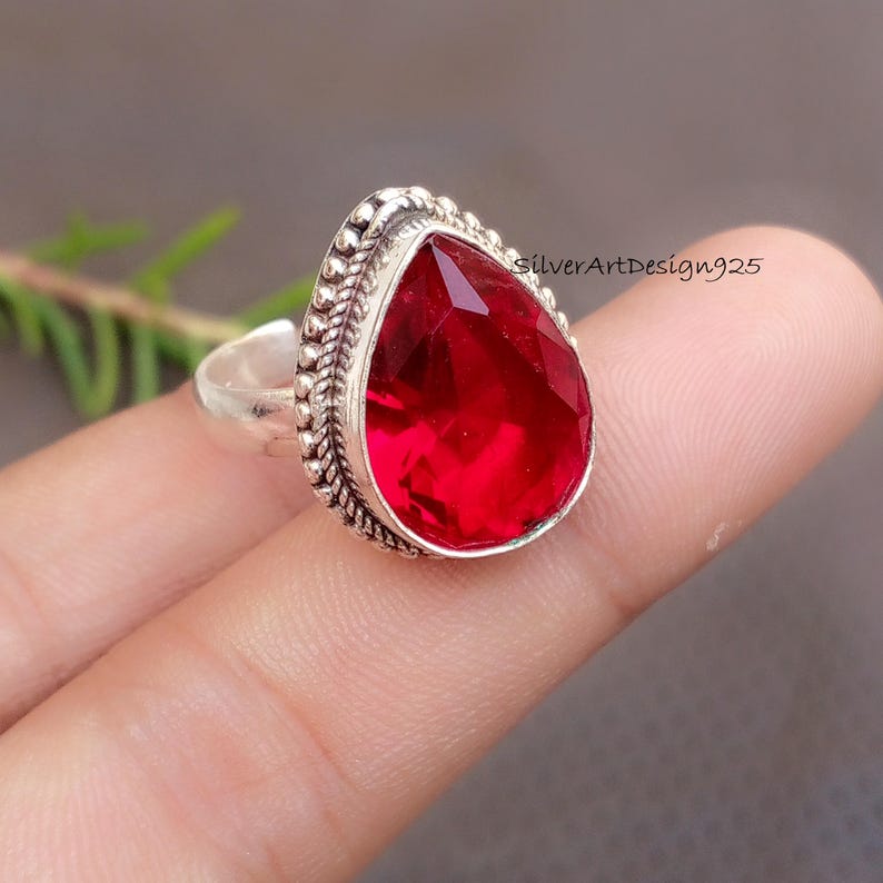 May include: A silver ring featuring a large, faceted, teardrop-shaped red gemstone. The gemstone is set in a decorative silver bezel with a beaded edge. The ring is displayed on a finger, with the text "silverArtDesign925" visible.