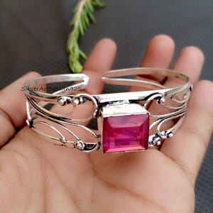 Può includere: Un bracciale a polsino in argento con un design aperto e una grande pietra preziosa rosa quadrata al centro. Il bracciale presenta intricati dettagli in filigrana e il testo "SilverArtDesign925".