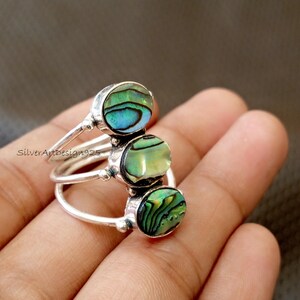 Pode incluir: Três anéis de prata, cada um com uma pedra redonda de concha de abalone. As pedras de abalone exibem tons verdes e azuis iridescentes. Os anéis são empilhados, criando um design único. Os anéis estão em uma mão.