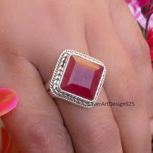 May include: A sterling silver ring featuring a large, square-cut ruby gemstone. The ring has a detailed, braided design around the ruby. The text "SilverArtDesign925" is visible on the ring.