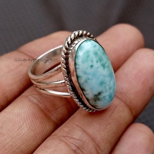 May include: A silver ring featuring a large, oval, pale blue gemstone with green marbling. The ring has a triple-band design and the gemstone is framed with a twisted rope-like border. The text "SilverArtDesign925" is visible. A piece of jewellery.