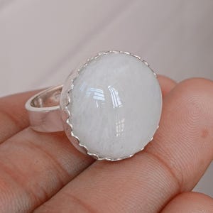 May include: A silver ring featuring a large, round, white gemstone. The gemstone has a smooth, polished surface and is set in a decorative, scalloped bezel. The ring's band is a simple, polished silver.