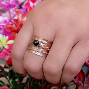 May include: A copper ring with a black gemstone and a textured design. The ring is made of multiple bands that are stacked on top of each other.