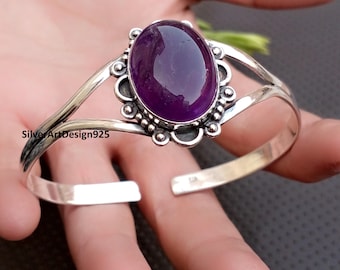 May include: A silver cuff bracelet featuring a large, oval, deep purple gemstone. The gemstone is set in a decorative bezel with a scalloped edge. The bracelet has an open design and is displayed on a hand.