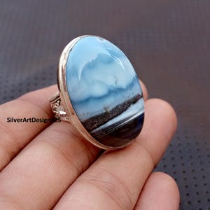 May include: An oval ring with a large gemstone in shades of blue, black, and white, set in a silver band with detailed patterns. The ring is held in a hand. The gemstone has a polished finish.