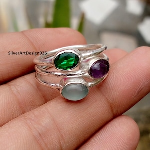 May include: A stackable silver ring set with three oval gemstones: green, purple, and light blue. The rings are displayed on a hand, showcasing the polished metal and vibrant colors of the stones.