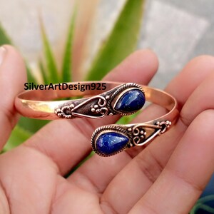 Puede incluir: Brazalete de puño cobrizo con detalles plateados y dos gemas azul oscuro en forma de lágrima. El brazalete abierto presenta el texto "SilverArtDesign925".