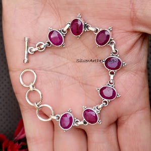Può includere: Un bracciale in argento con pietre preziose ovali rosso intenso. Il bracciale ha una chiusura a toggle ed è composto da maglie interconnesse. Ogni pietra preziosa è incastonata in una montatura d'argento decorativa.