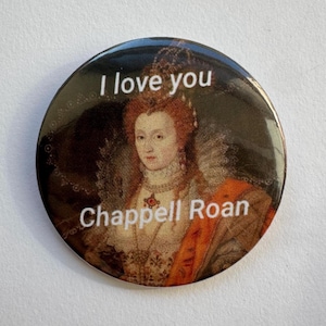 May include: A round button with a portrait of a woman with red hair wearing a gold and orange dress. The text "I love you" is at the top of the button and the text "Chappell Roan" is at the bottom.