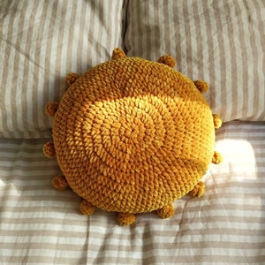 May include: A yellow crocheted sun cushion with a textured surface. The cushion is on a striped bedspread.