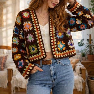May include: A brown, open-front crochet jacket with colorful floral squares. The jacket features a V-neck and long sleeves. The floral squares are in shades of blue, yellow, orange, and pink. The jacket is paired with blue jeans and a brown belt.