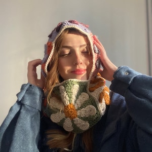 May include: A woman wearing a crocheted hooded scarf with a floral pattern in shades of green, orange, and white. The scarf is worn over a blue shirt.