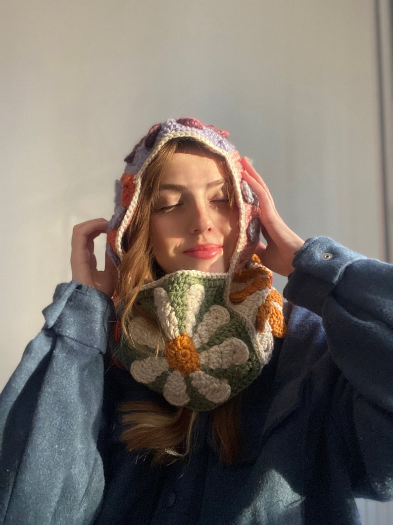 May include: A woman wearing a crocheted hooded scarf with a floral pattern in shades of green, orange, and white. The scarf is worn over a blue shirt.