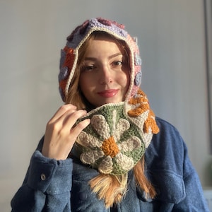 May include: A crocheted hat and scarf set with a floral design in shades of pink, orange, green, and white. The hat has a hood and the scarf is tied around the neck.