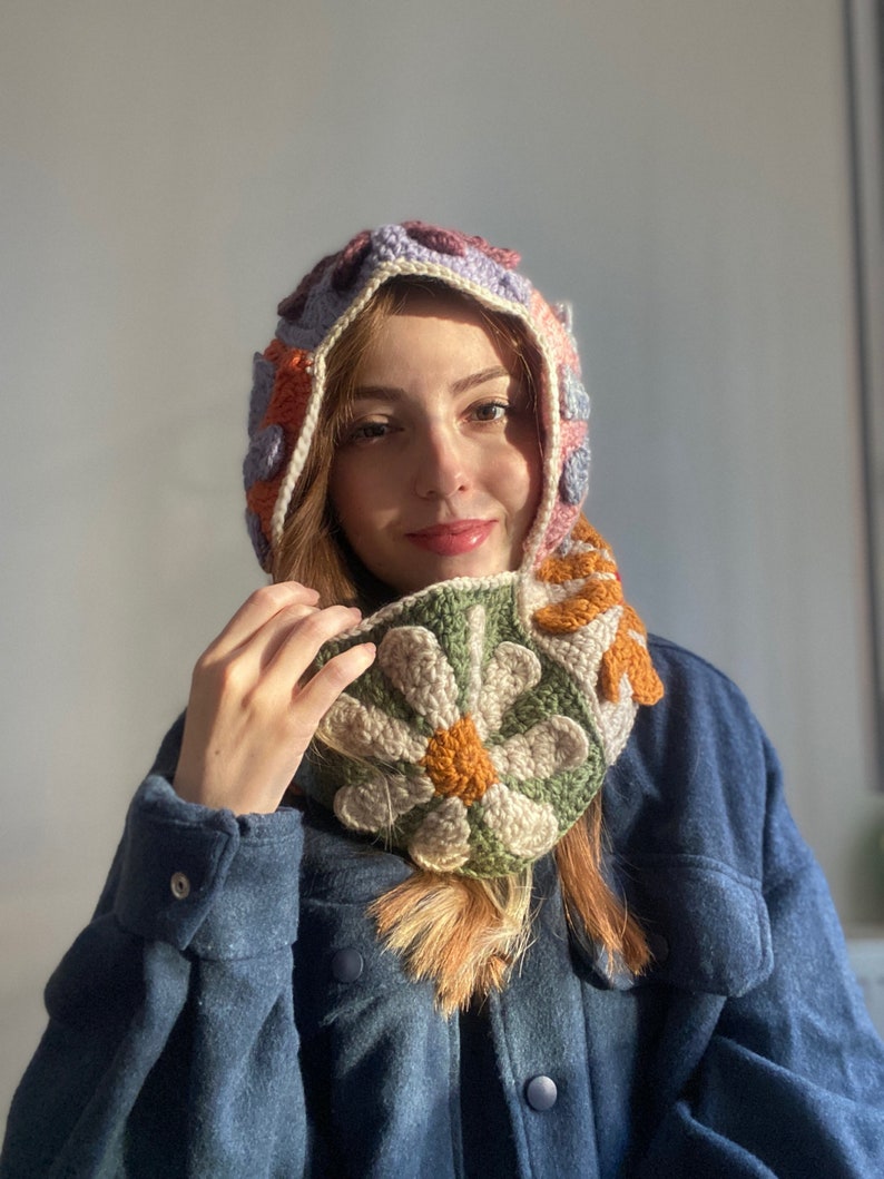 May include: A crocheted hat and scarf set with a floral design in shades of pink, orange, green, and white. The hat has a hood and the scarf is tied around the neck.