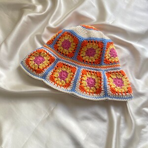 May include: A crocheted bucket hat with a white base and colorful granny squares in shades of orange, yellow, pink, and blue.