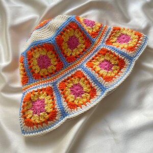 May include: A crocheted bucket hat with a white base and colorful granny squares in shades of orange, yellow, pink, and blue.