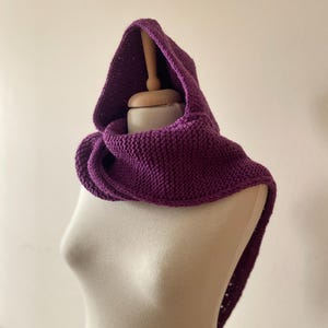 Puede incluir: Un cuello con capucha morado tejido, exhibido en un maniquí. El cuello es de un tono morado intenso, con un diseño de capucha y bufanda. La textura tejida es visible, lo que sugiere una prenda suave y cálida, ideal para el clima frío.
