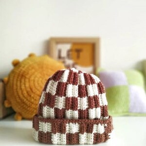 Może przedstawiać: Czapka beanie z dzianiny w brązowo-białe kratki. Czapka ma gruby, fakturowany materiał i podwinięty brzeg.