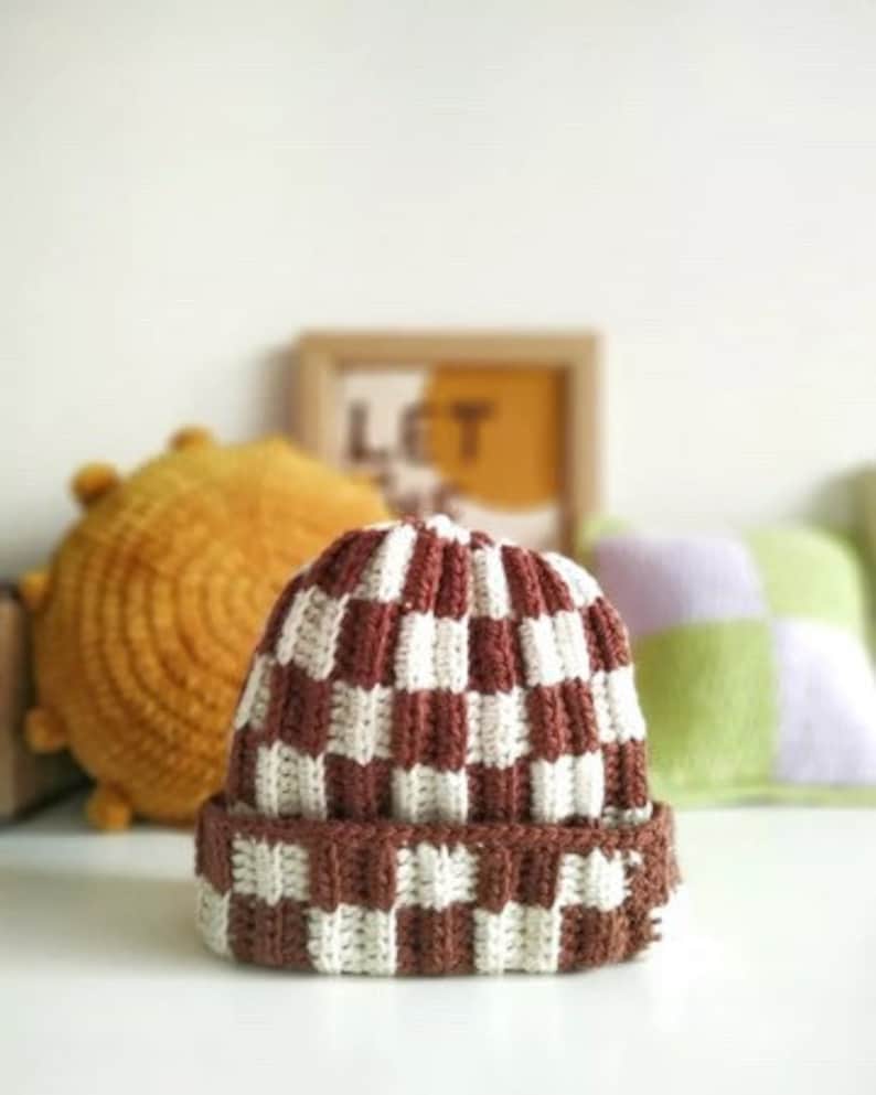 Może przedstawiać: Czapka beanie z dzianiny w brązowo-białe kratki. Czapka ma gruby, fakturowany materiał i podwinięty brzeg.