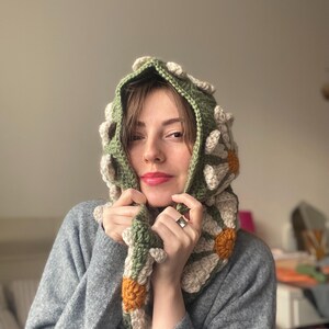 May include: A green crocheted hooded scarf with white and orange daisy flowers. The scarf is worn by a person with brown hair and is pulled up around their neck.