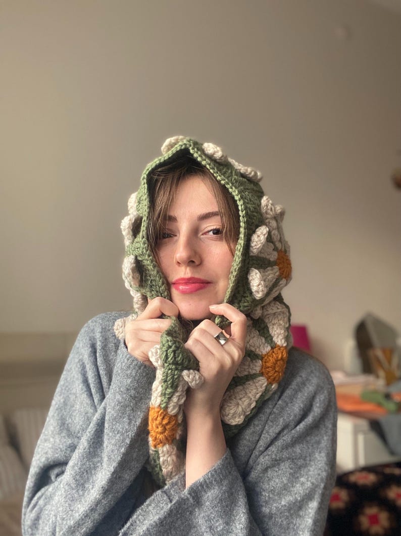 May include: A green crocheted hooded scarf with white and orange daisy flowers. The scarf is worn by a person with brown hair and is pulled up around their neck.