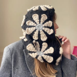 May include: A black crocheted hooded cowl with a white and gray floral design. The cowl is worn by a person with long brown hair.