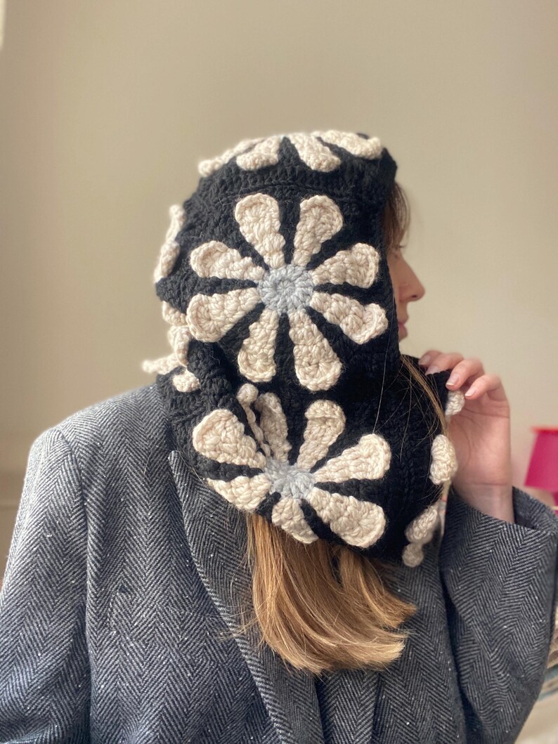 May include: A black crocheted hooded cowl with a white and gray floral design. The cowl is worn by a person with long brown hair.