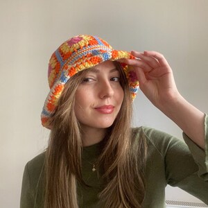 May include: A woman wearing a colorful crocheted bucket hat with orange, yellow, blue, and pink squares. The hat has a wide brim and is made with a chunky yarn.