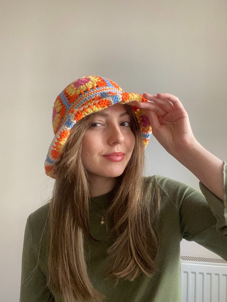 May include: A woman wearing a colorful crocheted bucket hat with orange, yellow, blue, and pink squares. The hat has a wide brim and is made with a chunky yarn.