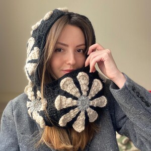 May include: A black and cream crocheted cowl with a large flower design. The cowl is worn by a person with long brown hair.