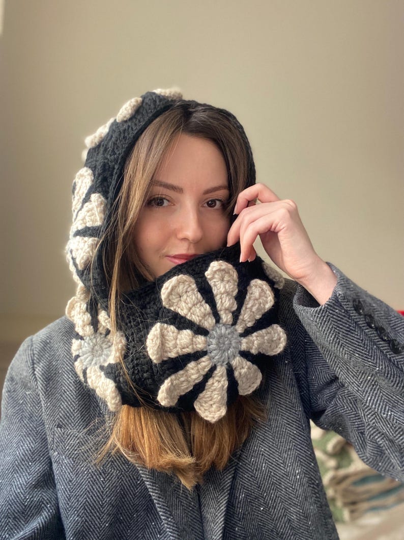 May include: A black and cream crocheted cowl with a large flower design. The cowl is worn by a person with long brown hair.