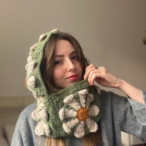 May include: A green crocheted hooded cowl with a large white daisy flower design. The flower has a yellow center and white petals.