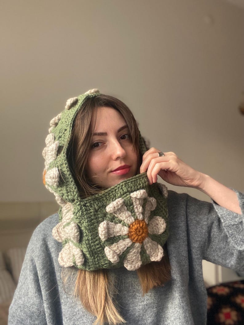 May include: A green crocheted hooded cowl with a large white daisy flower design. The flower has a yellow center and white petals.