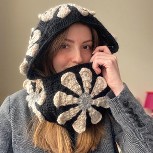 May include: A black and beige crocheted hooded scarf with a flower design. The scarf is worn by a person with brown hair.