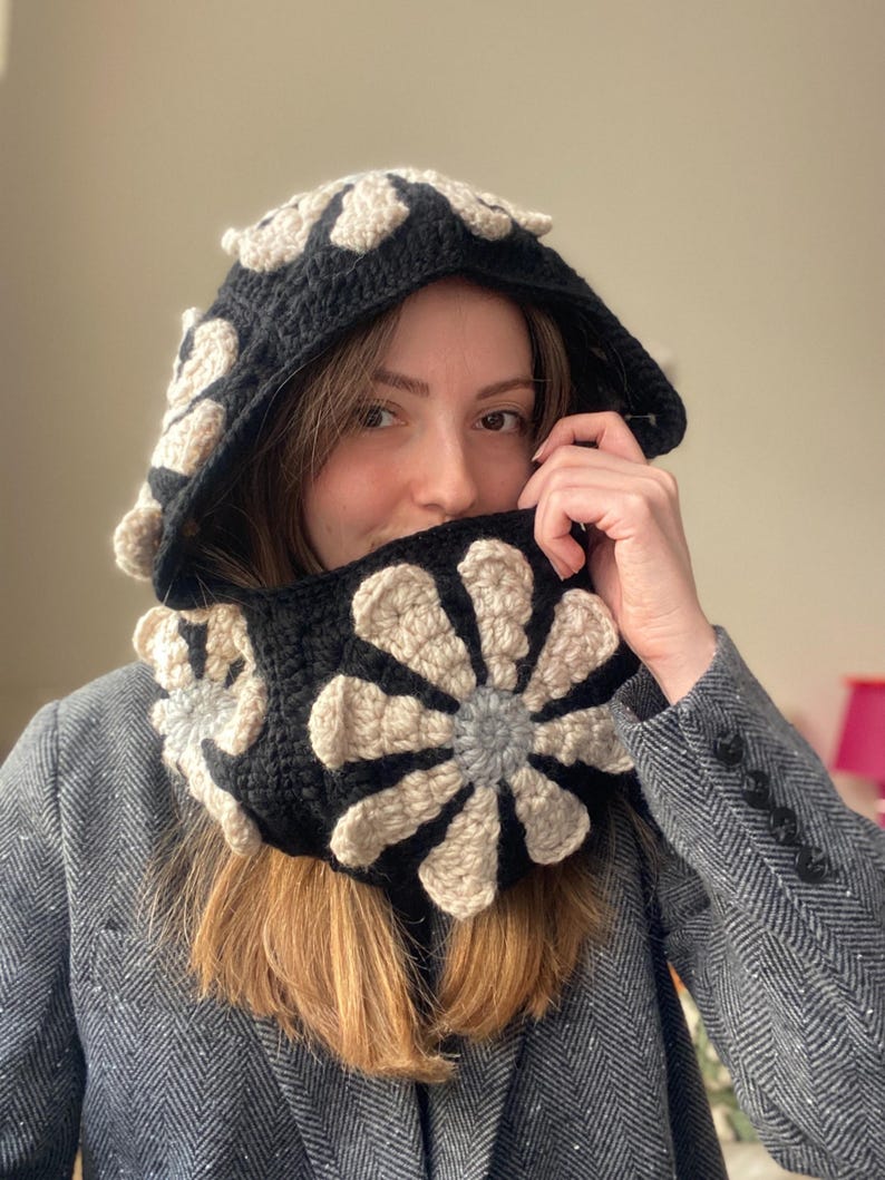 May include: A black and beige crocheted hooded scarf with a flower design. The scarf is worn by a person with brown hair.
