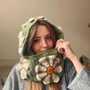 May include: A green crocheted hooded cowl with a large white daisy flower design. The flower has a yellow center.