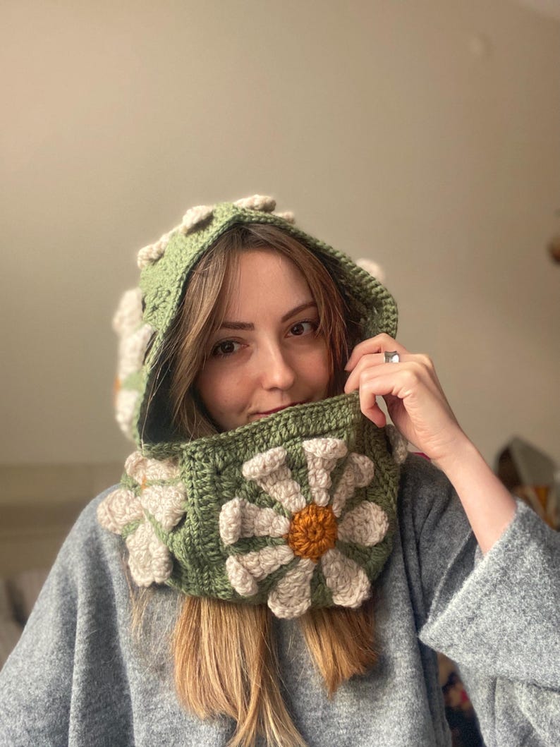 May include: A green crocheted hooded cowl with a large white daisy flower design. The flower has a yellow center.