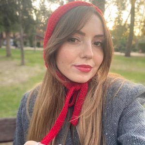 Puede incluir: Una mujer lleva un gorro de punto rojo con una larga bufanda atada alrededor del cuello. El gorro y la bufanda están hechos de un hilo suave y grueso.