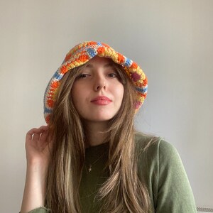 May include: A woman wearing a colorful crocheted bucket hat with a green long-sleeved shirt.