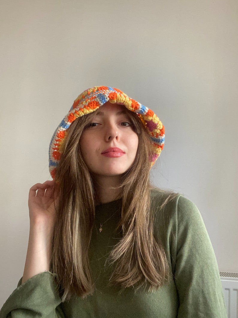 May include: A woman wearing a colorful crocheted bucket hat with a green long-sleeved shirt.