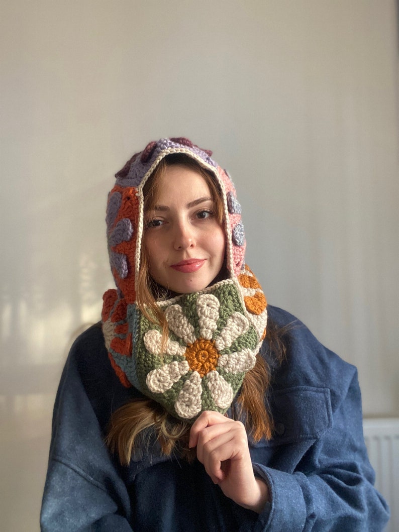 May include: A crocheted hooded scarf with a colorful geometric pattern and a large daisy flower design. The scarf is made with a variety of colors, including pink, orange, purple, green, and brown.