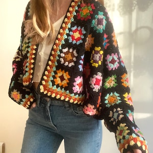 Puede incluir: Una chaqueta de punto de ganchillo negra con cuadrados de abuelita coloridos. La chaqueta tiene cuello en V y mangas largas. La chaqueta es corta y tiene un ajuste holgado.