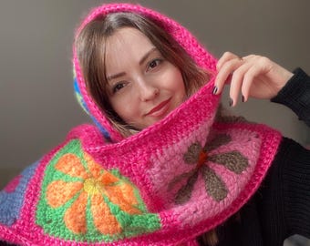 Peut inclure: Une écharpe en crochet rose avec une capuche, ornée de motifs floraux colorés. L'écharpe est réalisée avec un fil épais et a une texture douce et duveteuse.