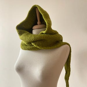 May include: A green knitted cowl with a long tail. The cowl has a hood that can be pulled up over the head.