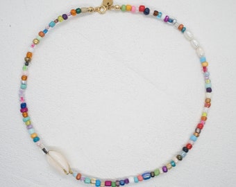 May include: A colorful beaded necklace with a shell charm. The necklace is made with a variety of small, colorful beads, including white, blue, green, red, orange, yellow, and purple. The necklace is finished with a gold clasp.